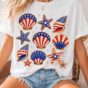 May include: White t-shirt featuring a patriotic design of seashells and starfish in red, white, and blue. The design includes gold accents and small diamond shapes. The shirt is a casual, everyday wear item.