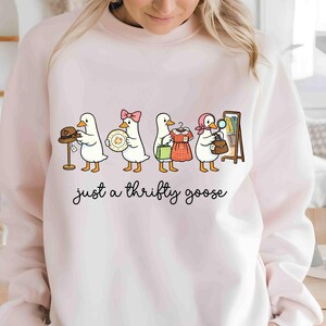 May include: A pale pink sweatshirt with a cartoon illustration of four geese, each holding a different item, such as a hat, plate, dress, and purse. The text "just a thrifty goose" is written below the geese in a cursive font.