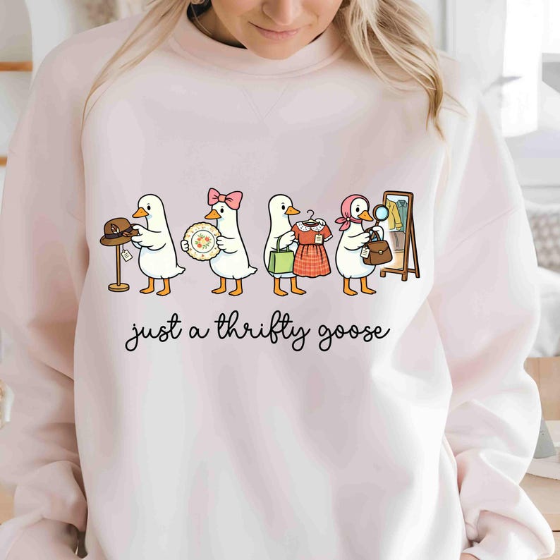 May include: A pale pink sweatshirt with a cartoon illustration of four geese, each holding a different item, such as a hat, plate, dress, and purse. The text "just a thrifty goose" is written below the geese in a cursive font.