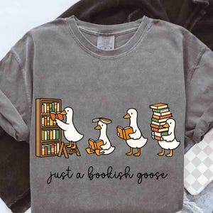 May include: Gray sweatshirt featuring a whimsical illustration of four cartoon geese with books. One goose is reading from a bookshelf, another is reading while sitting, and the others are holding stacks of books. The text "just a bookish goose" is below.