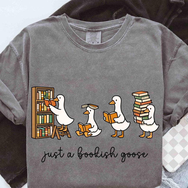 May include: Gray sweatshirt featuring a whimsical illustration of four cartoon geese with books. One goose is reading from a bookshelf, another is reading while sitting, and the others are holding stacks of books. The text "just a bookish goose" is below.