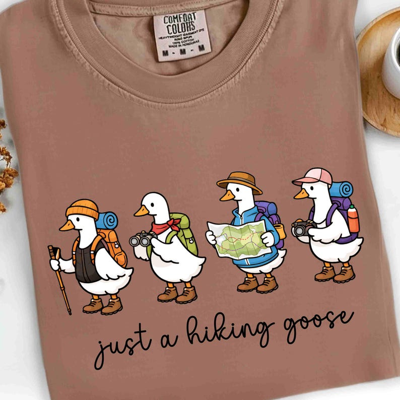 K&ouml;nnte beinhalten: Ein braunes Comfort Colors T-Shirt mit einer Illustration von vier Cartoon-G&auml;nsen, die zum Wandern bereit sind. Jede Gans tr&auml;gt einen Rucksack und verschiedene Wanderaccessoires. Der Text "just a hiking goose" steht in Kursivschrift darunter.