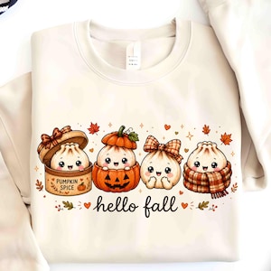 May include: A cream-colored crewneck sweatshirt featuring a graphic of four cute, smiling dumpling characters. One is in a pumpkin spice box, one in a jack-o'-lantern, one with a plaid bow, and one in a plaid scarf. The text reads "hello fall".