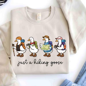 K&ouml;nnte beinhalten: Beiges Sweatshirt mit einer skurrilen Illustration von vier Comic-G&auml;nsen, die sich auf eine Wanderung vorbereiten, jede mit unterschiedlichem Zubeh&ouml;r wie Rucksack, Karte und Kamera. Der Text "just a hiking goose" steht darunter in Schreibschrift.