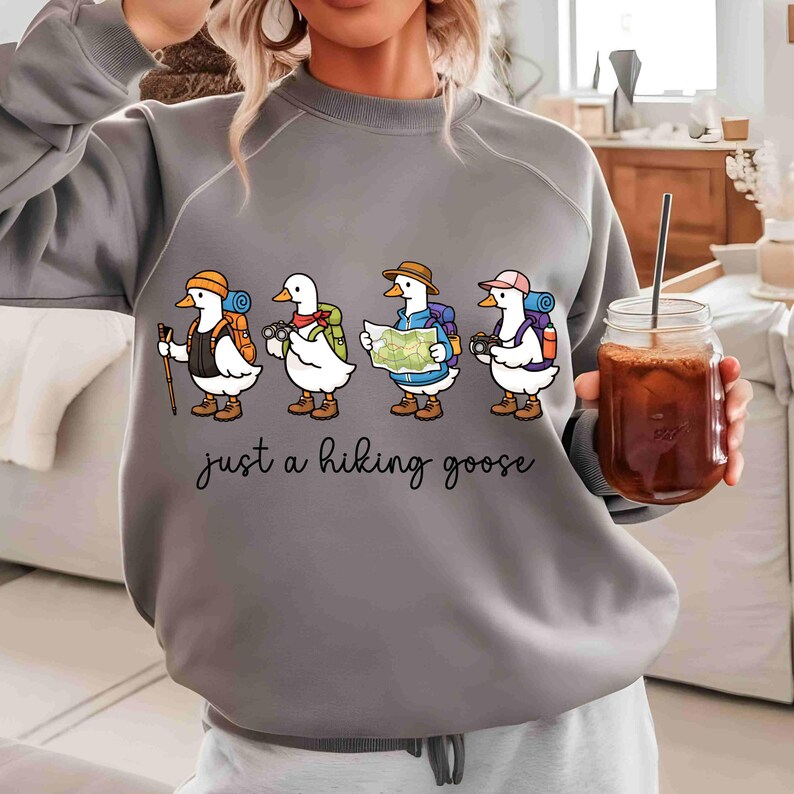 K&ouml;nnte beinhalten: Graues Sweatshirt mit einem skurrilen Design von vier Cartoon-G&auml;nsen, die zum Wandern gekleidet sind, jede mit Rucksack und Ausr&uuml;stung. Der Satz "just a hiking goose" ist darunter in Kursivschrift geschrieben.