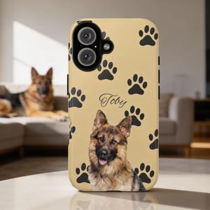Op de afbeelding: Een beige telefoonhoesje met een portret van een Duitse herdershond, zwarte pootafdrukken en de naam "Toby". De hoes heeft een uitsparing voor een camera met dubbele lens en een zwarte rand. Op de achtergrond is een Duitse herdershond te zien.