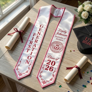 May include: White graduation sash with pink edging and embroidered text. One sash reads "GENERATION FIRST" and the other has a name, degree, and "Class of 2026." A graduation cap and diploma are also present.