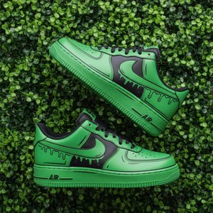 May include: A pair of green and black trainers with a distinctive design. The shoes have a green base with black detailing, including the Nike swoosh and a dripping paint effect. The laces and soles are green.