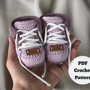 May include: A pair of handmade crochet baby sneakers in shades of lavender and mauve, featuring white laces and small brown tags. The sneakers are designed to resemble adult athletic shoes. The image also includes a white circle with the text "PDF Crochet Pattern".
