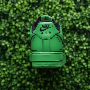 May include: Green and black Nike Air Force 1 trainers. The heel displays the Nike Air logo in black on a green panel. Black dripping paint design on the sides. The trainers are set against a green background.