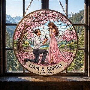 May include: A circular stained glass suncatcher featuring a proposal scene with a person kneeling to offer a ring to another person. The design includes cherry blossom trees, a lake, and a banner with the text LIAM & SOPHIA - EST. 2024.