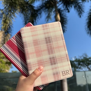 May include: Two notebooks, one with a red and white striped pattern and the other with a pink and beige plaid design. The plaid notebook has the initials "BR" in the corner. The background is a bright blue sky and palm trees.