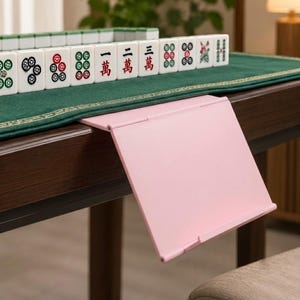 May include: A light pink plastic folding stand attached to the edge of a dark wooden table. A green felt mat covers the table surface, featuring a row of white mahjong tiles with various red, green, and black symbols and Chinese characters.