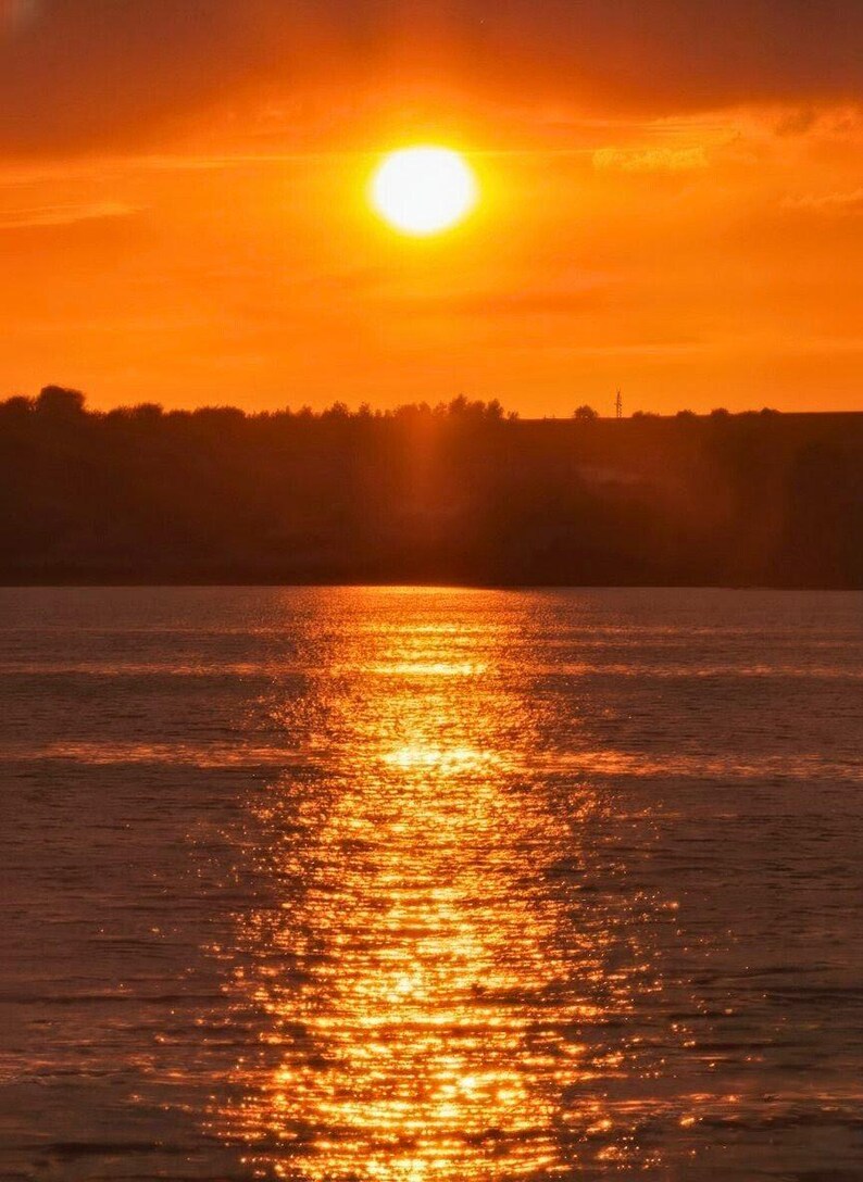 May include: A vivid sunset scene over water. The sun, a bright disc, reflects a golden light across the water. The sky is a gradient of orange and yellow, with a dark silhouette of trees and land in the distance.