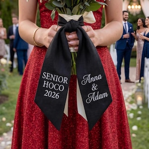 Könnte beinhalten: Ein Blumenstrauß ist mit einem schwarzen Band gebunden. Das Band trägt auf der einen Seite den weißen Text "SENIOR HOCO 2026" und auf der anderen Seite "Anna & Adam". Die Person, die den Strauß hält, trägt ein rotes Paillettenkleid.