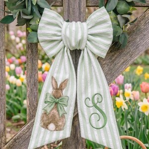 Puede incluir: Un gran lazo decorativo con rayas verdes y blancas, con un conejito marrón con un lazo verde y la letra "S" bordada en verde. El lazo cuelga de una valla de madera con un fondo floral.