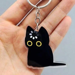 May include: A black cat-shaped keychain with a silver keyring and chain. The cat has yellow eyes and white and yellow accents on its head. The keychain is held in a hand.