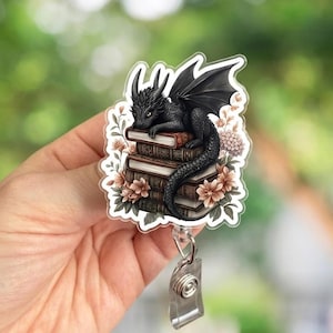 May include: A badge reel featuring a black dragon resting on a stack of books, surrounded by flowers. The dragon has detailed wings and horns. The reel has a clear plastic ID holder and a metal clip.