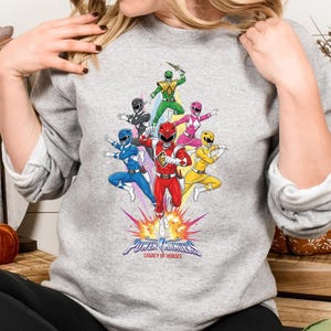 Könnte beinhalten: Ein graues Sweatshirt mit einer Grafik der Power Rangers in Action. Die Illustration zeigt die roten, blauen, schwarzen, grünen, pinken und gelben Rangers. Der Text "Power Rangers Legacy of Heroes" ist unter den Charakteren zu sehen.