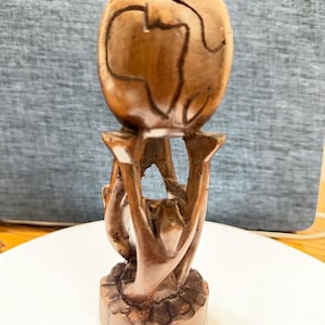 May include: A hand-carved wooden sculpture featuring a spherical globe with a map of the African continent etched into the surface. The globe is supported by a base consisting of three stylised figures standing on a circular, textured pedestal.