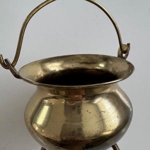 Puede incluir: Un pequeño caldero de color latón con un asa curva y tres patas pequeñas. El caldero tiene un cuerpo redondeado y un borde ancho. El asa está sujeta a los lados. El interior es oscuro y el exterior tiene un aspecto ligeramente envejecido.