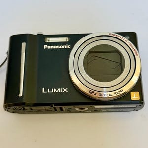 May include: A black Panasonic Lumix digital camera featuring a 12x optical zoom lens with Power O.I.S. and 25mm wide capabilities. The front of the device displays the Panasonic and Lumix brand names, a flash unit, and a gold L logo.