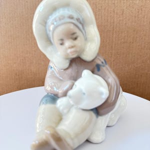 May include: A glossy porcelain figurine depicting a child wearing a hooded winter parka with a fur-trimmed hood, sitting down and embracing a small white polar bear cub. The figure features soft brown, cream, and light blue painted accents.