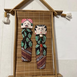Peut inclure: Décoration murale en bois avec deux figurines sculptées en costume traditionnel. Les figurines sont placées sur un tapis en bambou, encadrées par une structure en bois avec des pompons. Les figurines ont des visages peints et des vêtements détaillés.
