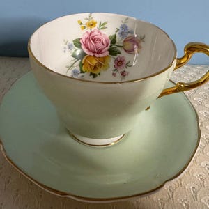 Puede incluir: Un juego de taza y platillo de té vintage. La taza es blanca con un diseño floral con rosas rosas y amarillas. El asa es dorada. El platillo y el exterior de la taza son de color verde pálido. Los bordes son dorados.