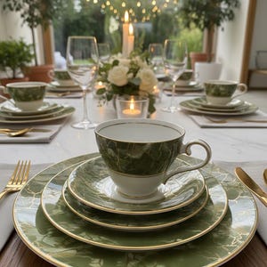 Puede incluir: Elegante mesa puesta con porcelana verde y dorada. El conjunto incluye tazas, platillos y platos, dispuestos con cubiertos dorados, copas de vino y velas. Un centro de mesa floral añade un toque de sofisticación a la disposición.