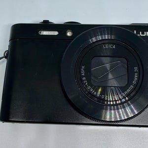 May include: A black Panasonic Lumix digital camera featuring a Leica DC Vario-Summilux lens with 1:2.0-5.9/16.0-42.8 ASPH specifications. The camera body includes a top-mounted flash, a small L logo on the front, and a grey wrist strap with Lumix branding.