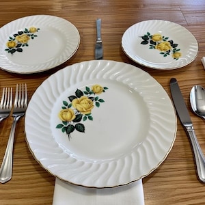 May include: A white porcelain dinner set featuring a yellow rose floral pattern. The set includes plates, forks, knives, and spoons. The plates have a gold trim and a ridged edge. The table setting is on a wooden surface.
