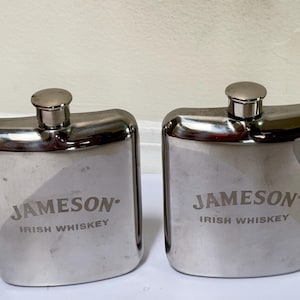 Puede incluir: Dos petacas de metal plateado con esquinas redondeadas y tapones de rosca. Cada petaca presenta el texto grabado "JAMESON" sobre las palabras "IRISH WHISKEY" centradas en la parte frontal. El acabado pulido refleja la luz sobre los cuerpos curvos.