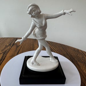 May include: A white porcelain figurine of a figure skater in a graceful pose. The sculpture is mounted on a black rectangular base. The skater is wearing a short skirt and a hat. The figurine is on a white circular surface.