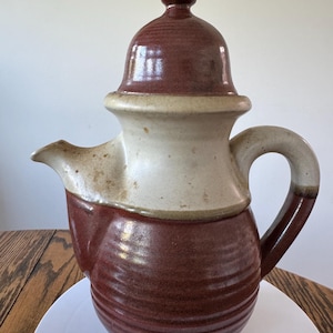 Pode incluir: Uma chaleira de cerâmica artesanal com tampa. A chaleira apresenta uma base e tampa vermelha escura, uma seção intermediária creme e uma alça curva. O bico está no lado esquerdo. A chaleira está em um prato branco.