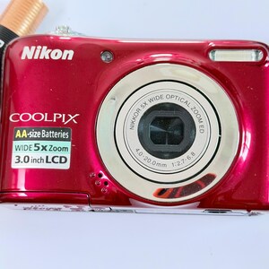 May include: A metallic red Nikon Coolpix digital camera with a silver lens ring. The front features a sticker stating AA-size Batteries, WIDE 5x Zoom, and 3.0 inch LCD. A black wrist strap and a Duracell AA battery are positioned next to the camera.