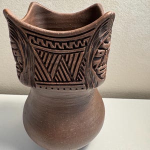 Puede incluir: Un jarrón de cerámica marrón con un borde ancho y decorativo y una base bulbosa. El jarrón presenta patrones geométricos tallados y caras estilizadas alrededor del borde. El jarrón mide aproximadamente 18 cm de alto.