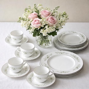 Peut inclure: Service à thé en porcelaine blanche avec un motif floral et un liseré argenté, comprenant tasses, soucoupes et assiettes. Un bouquet de roses roses et de fleurs blanches est dans un vase en verre transparent.