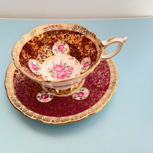 Könnte beinhalten: Ein Porzellan-Tassenset mit Untertasse in tiefem Burgunderrot, verziert mit filigranen Goldmustern. Das Innere der Tasse und die Untertasse sind mit weißen, kreisförmigen Medaillons versehen, die handgemalte rosa Rosensträuße zeigen.