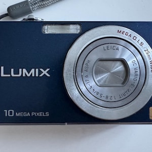 May include: A navy blue Lumix digital camera with a Leica lens. The camera has a silver lens ring and a built-in flash. The text "LUMIX" and "10 MEGA PIXELS" are visible on the camera body. The camera has a gray wrist strap.