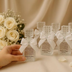 Puede incluir: Frascos de perfume de vidrio transparente con tapones cuadrados y lazos blancos. Cada botella tiene una etiqueta con texto. Un ramo de rosas blancas y una vela encendida están en el fondo.