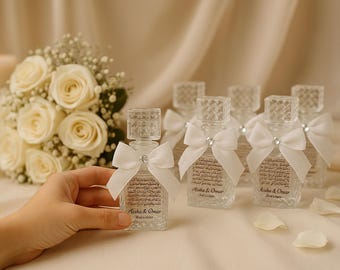 May include: Clear glass perfume bottles with square stoppers and white bows. Each bottle has a label with text. A bouquet of white roses and a lit candle are in the background. The bottles are on a white surface.