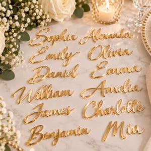 Può includere: Una collezione di segnaposto in acrilico color oro con vari nomi scritti in un carattere corsivo. I nomi includono Sophia, Alexander, Emily, Olivia, Daniel, Emma, William, Amelia, James, Charlotte, Benjamin e Mia.