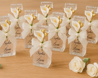 May include: Clear glass favor boxes decorated with white calla lilies, ivory bows, and the names "Sophia" and "Lucas" along with the date "27.01.2027". The favors are arranged on a wooden surface, with white roses and a decorative ball.