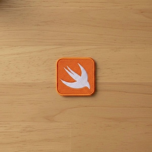 May include: Square orange patch with a white bird design. The patch is embroidered with a white bird in flight, set against a bright orange background. The patch is likely for clothing or accessories.