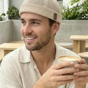 Op de afbeelding: Een beige, mesh-gebreide beanie met een kleine bruine patch. De persoon houdt een kopje koffie met latte art vast. De achtergrond bevat een bord met de tekst "Boon's Coffee".
