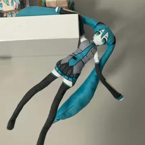 May include: A plush doll of a character with turquoise hair and eyes, dressed in a grey and teal outfit. The doll has long, black legs and arms, and is posed as if hanging from a shelf. Other papercraft figures are visible in the background.