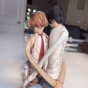 Peut inclure: Une sculpture en tissu de deux personnages s'embrassant. L'un a les cheveux bruns et une cravate rouge, l'autre a les cheveux noirs. Ils portent des chemises claires et des jeans bleus. La sculpture est sur une surface blanche.