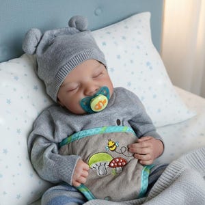 Op de afbeelding: Een slapende baby met een grijze gebreide muts met berenoren en een grijze trui. De baby heeft een fopspeen met een visontwerp. De baby draagt een luier met een geborduurd paddenstoel- en bijenontwerp. De baby ligt op een bed met een kussen met sterrenpatroon.