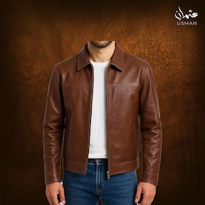 Puede incluir: Chaqueta de cuero marrón con cuello y cierre de cremallera. La chaqueta tiene un diseño clásico con costuras y bolsillos visibles. El modelo lleva una camiseta blanca y vaqueros azules. La marca "USMAN" es visible en la esquina superior derecha.